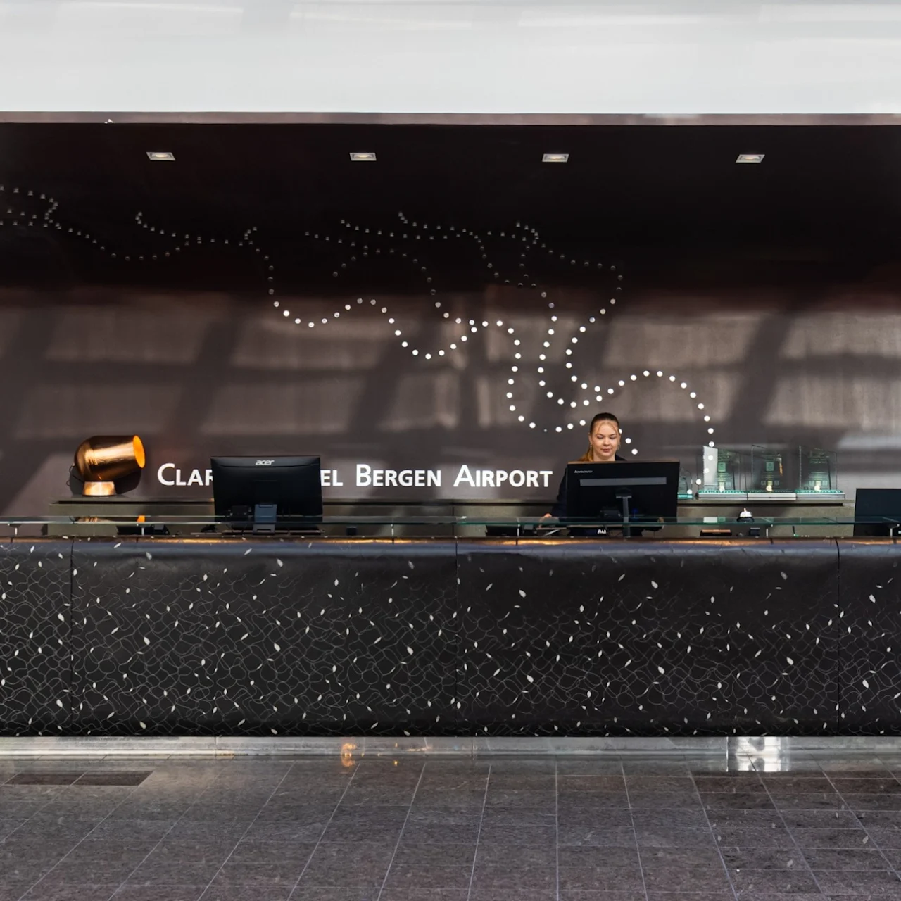 A long reception desk with computers and a staff member stands in a modern lobby. Behind, a black wall features dotted artwork and the text "CLARION HOTEL BERGEN AIRPORT."