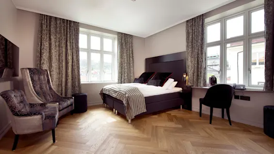 A bed with striped blanket and cushions stands in a modern room with large windows, wooden floors, and velvet chairs. A phone and coffee machine are on the windowsill.