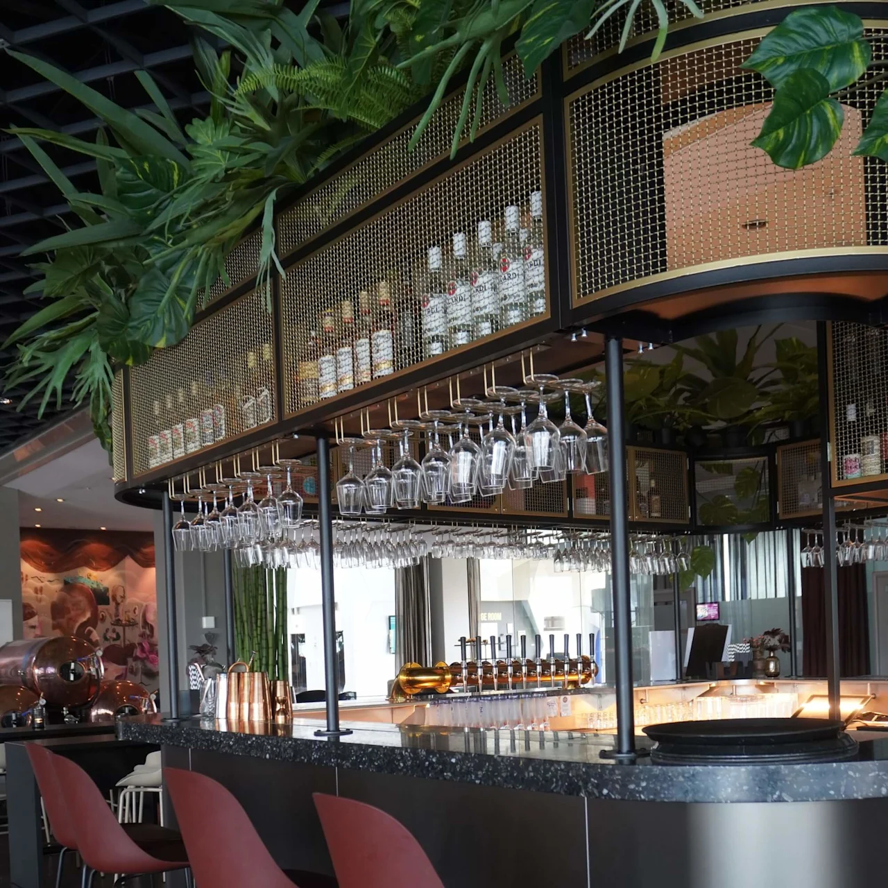 A modern bar with high chairs lines a sleek counter, showcasing hanging glassware and assorted bottles. Lush green plants adorn the overhead shelving, creating a stylish, inviting atmosphere.