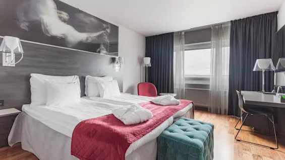A comfortable hotel room with two beds, white bathrobes, a teal ottoman, and a desk by a window with curtains.
