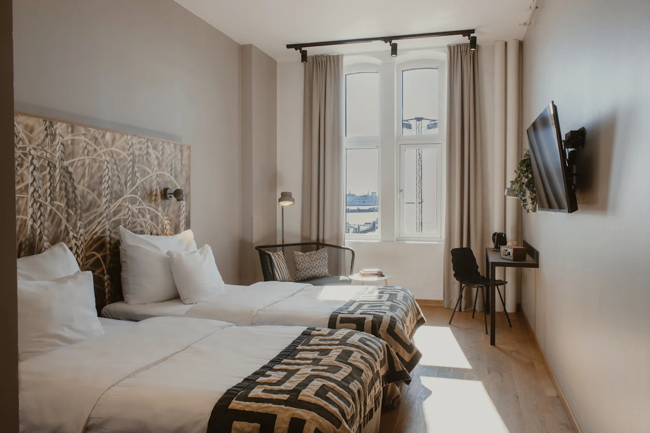 Valoisa hotellihuone, jossa kaksi erillistä sänkyä, kuvioitu sängynpääty, ikkuna näköalalla ja pieni työpöytäalue.