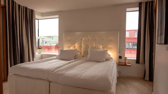 Ett mysigt hotellrum med två sängar, en upplyst sänggavel och stora fönster med utsikt över staden.