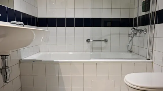 Badkar med en glasskärm och monterade kontrollhandtag, placerat mot vita och marinblå kakelväggar. Ett handfat och en toalett finns i förgrunden. Ingen text finns.