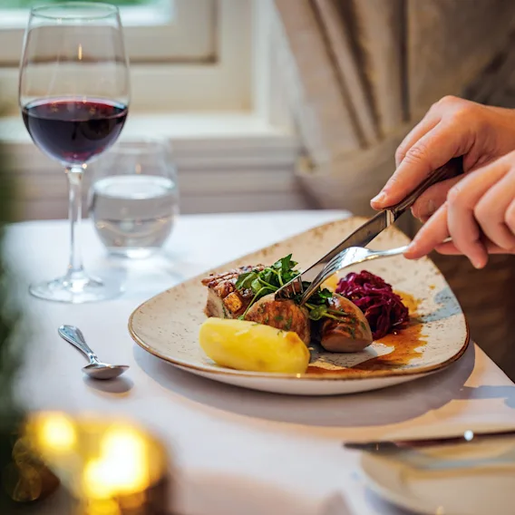 Nærbilde av hender som skjærer i en julerett ved et restaurantbord ved siden av et glass rødvin.