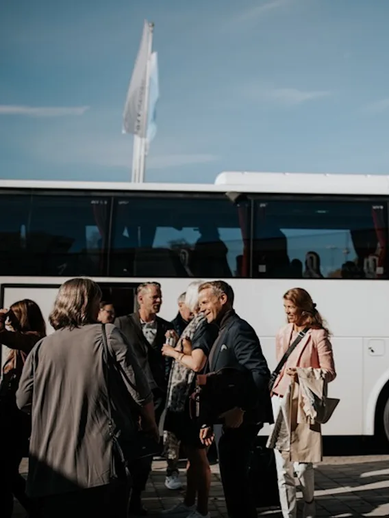 En grupp människor som pratar och ler utanför en vit buss under en klarblå himmel, möjligen före eller efter en resa.