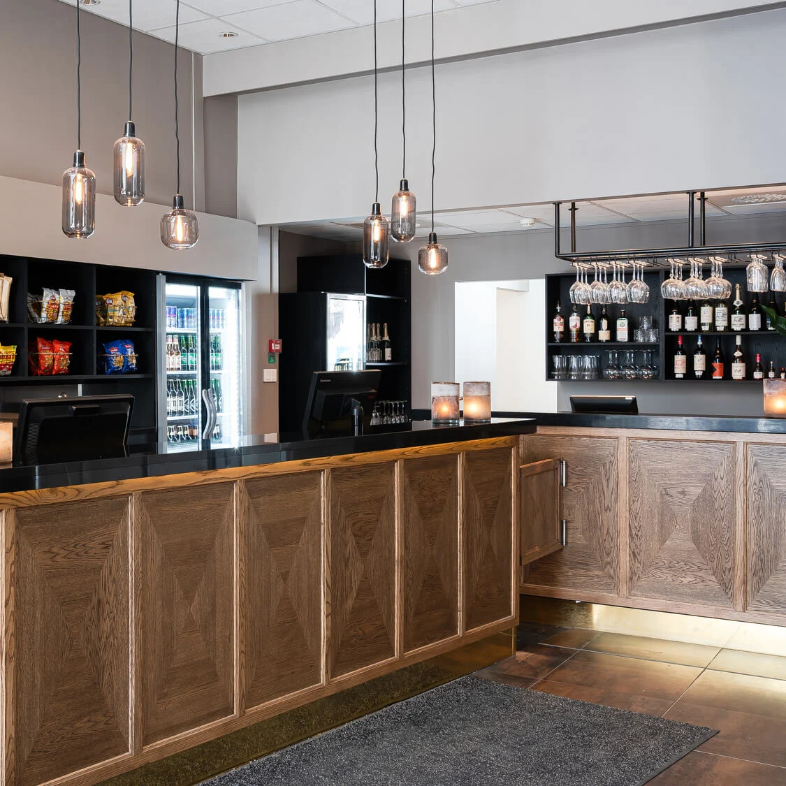 Disk som visar snacks i en modern hotellobby; belyst av hängande lampor, med träpaneler och dekorativa växter. En bardel med glas och flaskor i bakgrunden.