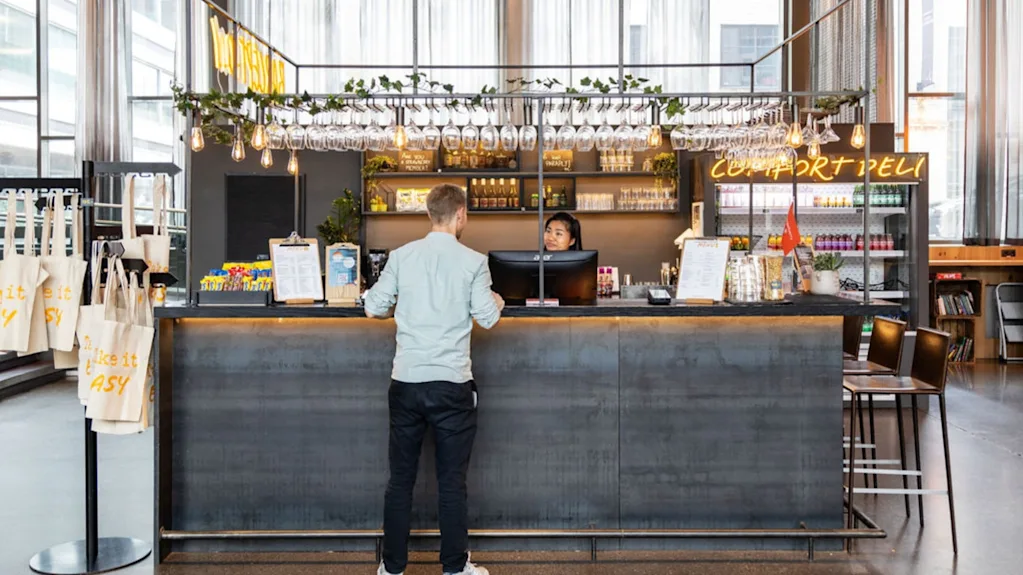 En mand står ved en moderne cafédisk og taler med personalet. Disken er dekoreret med hængende planter og glas; neonskiltet lyder "COMFORT DELI."