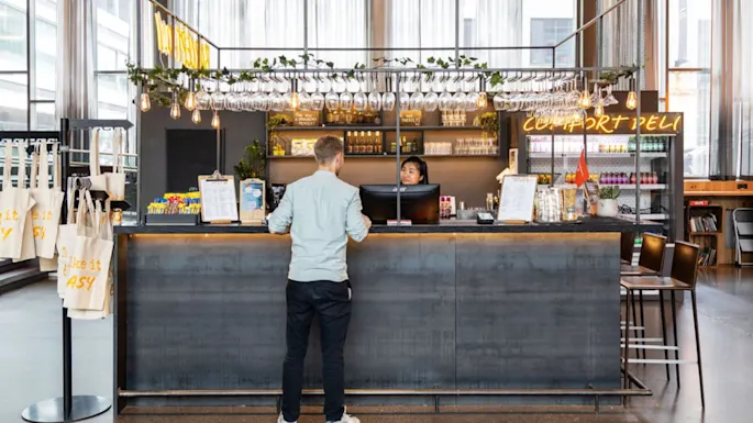 Man stands at counter in a modern café, speaking with a staff member. The counter is decorated with hanging plants and glasses; neon sign reads "COMFORT DELI."