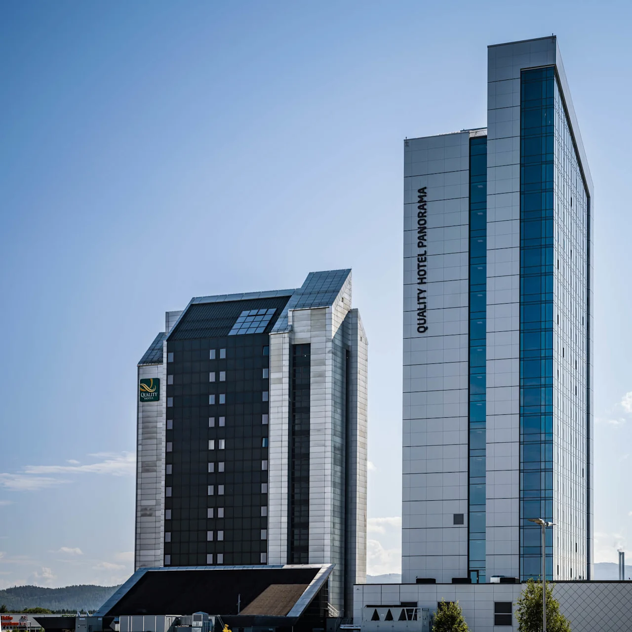 To høye bygninger står nær hverandre under en klarblå himmel. En har teksten "QUALITY HOTEL PANORAMA" vertikalt. Den urbane settingen har lave åser i bakgrunnen.