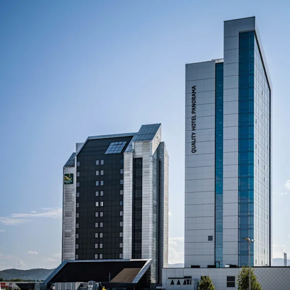 To høye bygninger står nær hverandre under en klarblå himmel. En har teksten "QUALITY HOTEL PANORAMA" vertikalt. Den urbane settingen har lave åser i bakgrunnen.