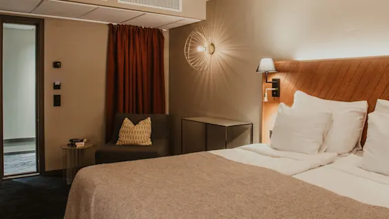 Viihtyisä hotellihuone mukavalla sängyllä ja lukunurkkauksella, lämpimällä valaistuksella, kutsuen vieraat rentoutumaan.
