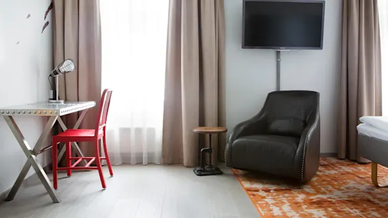 A desk with a red chair and lamp sits by a window with beige curtains. A black armchair and TV are against the wall.