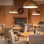 Tables and chairs arranged in a cozy cafe setting, illuminated by dome-shaped pendant lights. The warm, brown walls create an inviting ambiance with a decorative wreath above a fireplace.