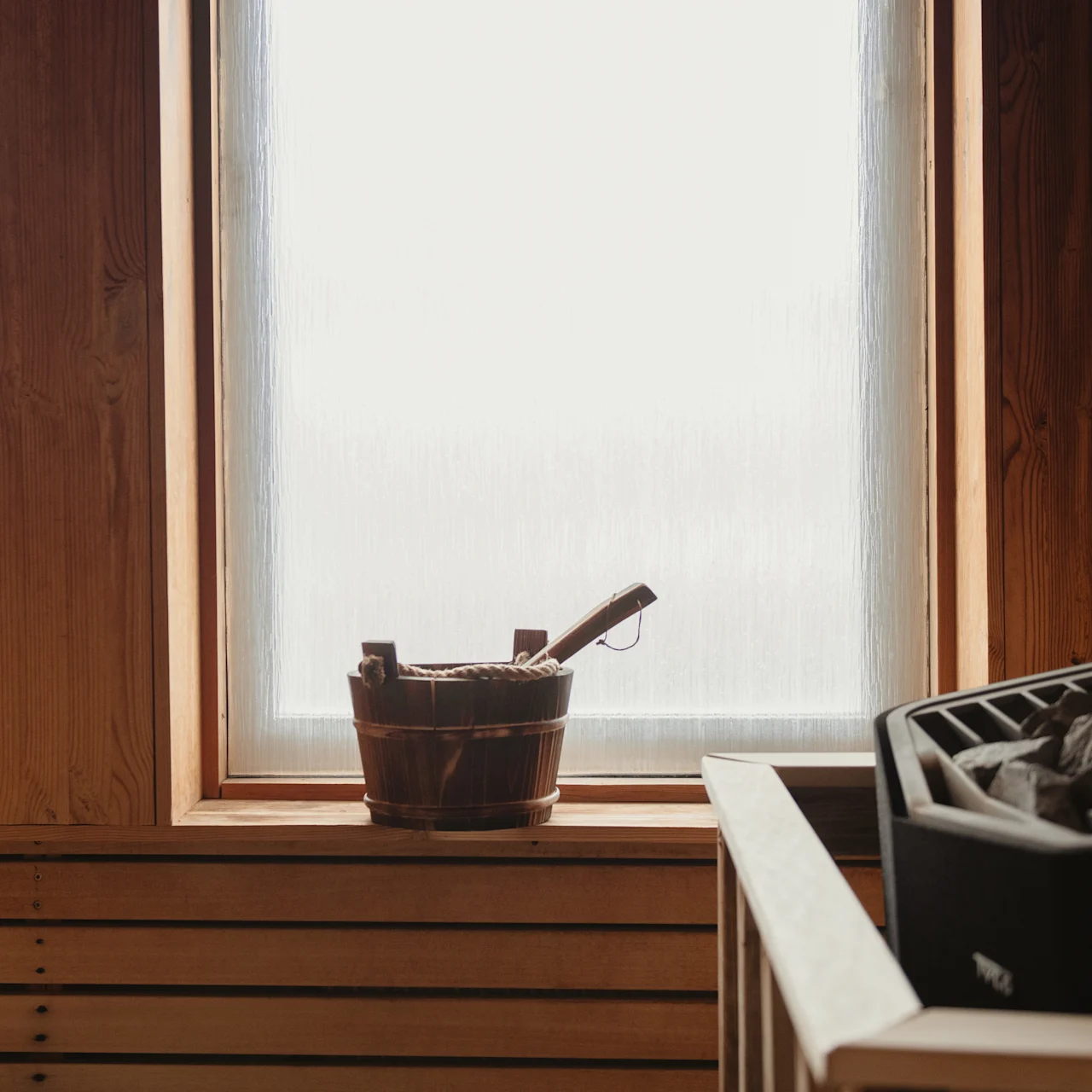 A wooden bucket sits on a windowsill, bathed in light, in a wooden sauna room with a heater filled with stones nearby.