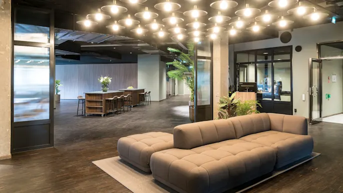 En romslig, moderne lounge med en brun sofa under lyse taklamper. I nærheten står et høyt bord med krakker og planter som skaper en innbydende atmosfære i et elegant kontormiljø.