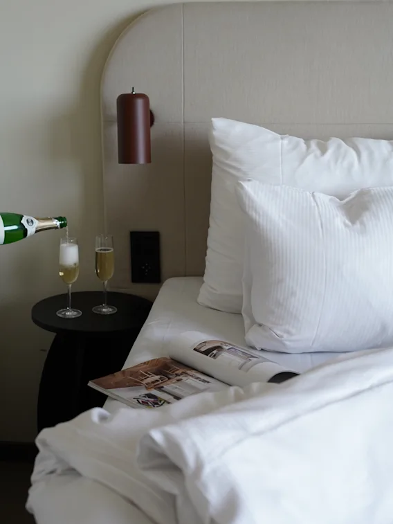 Champagne hälls upp i glas bredvid en bäddad hotellsäng med vita lakan och en tidning.