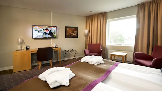 A hotel room with a made bed, towels on the floor, furniture, and a mounted television showing a man in a blurred background.