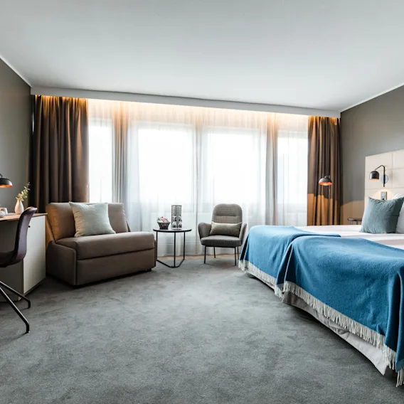 Ett rymligt hotellrum har en säng med blå och vita sängkläder, ett skrivbord med stol och laptop samt en sittgrupp vid stora fönster med gardiner.
