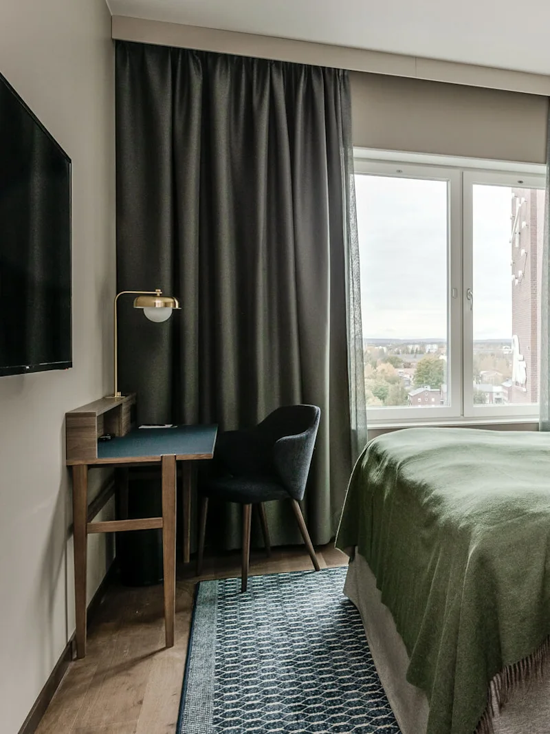 Huolellisesti pedattu sänky vihreällä peitolla sijaitsee ikkunan vieressä. Seinälle asennettu TV ja pieni pöytä tuolin kanssa ovat modernissa huoneessa.