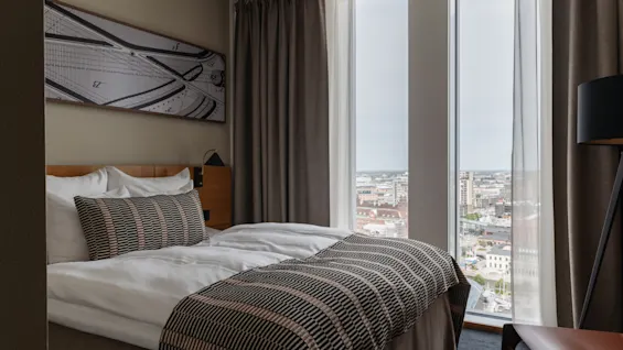 Et koselig hotellrom med en komfortabel seng, utsikt over byen fra et stort vindu og moderne innredning.