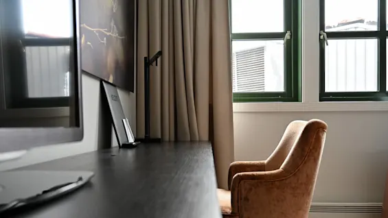 En plyschbrun fåtölj står vid ett svart skrivbord under två stängda fönster med gröna ramar. Beige gardiner hänger bredvid fönstren. En suddig datorskärm syns på skrivbordet.