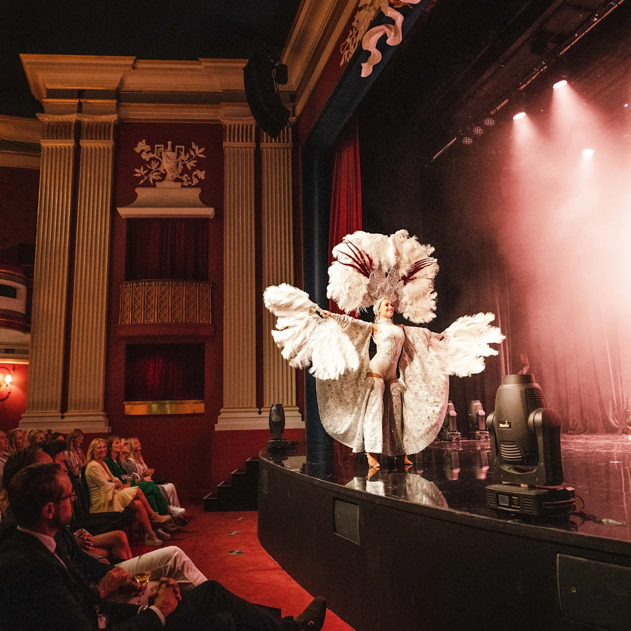 A performer wearing an elaborate feather costume dances on a stage, surrounded by elegant theater interiors, as an attentive audience watches from red velvet seats.