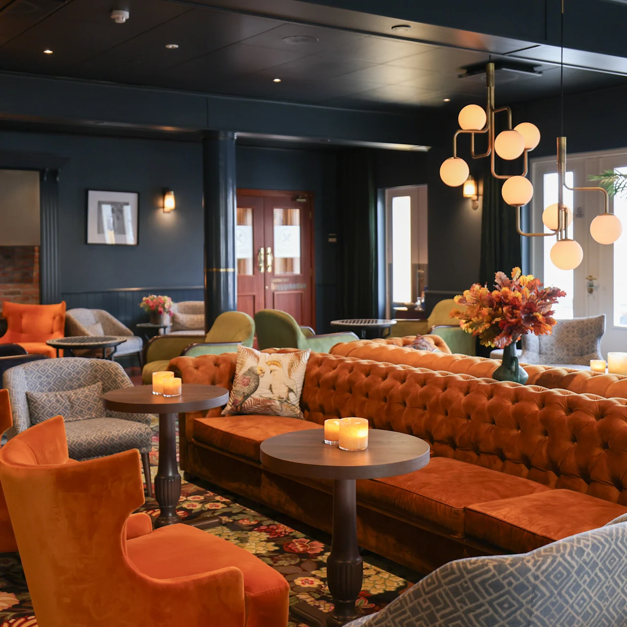 Luxurious lounge with orange velvet chairs surrounding wooden tables, featuring dim lighting from spherical chandeliers. Soft carpets, floral arrangements, and a warm fireplace create a cozy, inviting atmosphere.