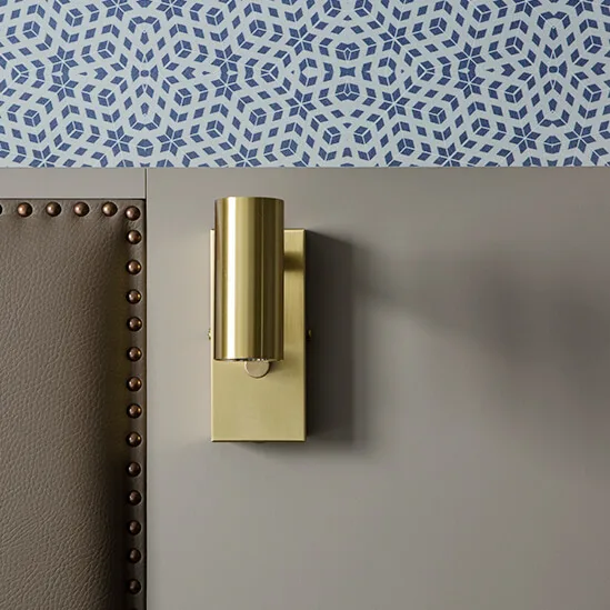 A gold wall sconce casts a shadow on a neutral wall above a studded couch, against patterned wallpaper.