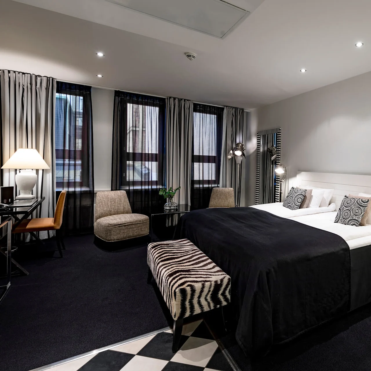 Et pent innredet hotellrom har en stor seng med svart sengetøy, mønstrete puter og en zebra-stripet benk. Moderne møbler og dekorasjoner skaper en stilig atmosfære.