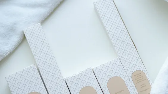 Boxes labeled "Drawing Kit" and "Serene Kit" are arranged on a white surface. Textured and patterned packaging is surrounded by soft white fabric, adding a cozy feel.