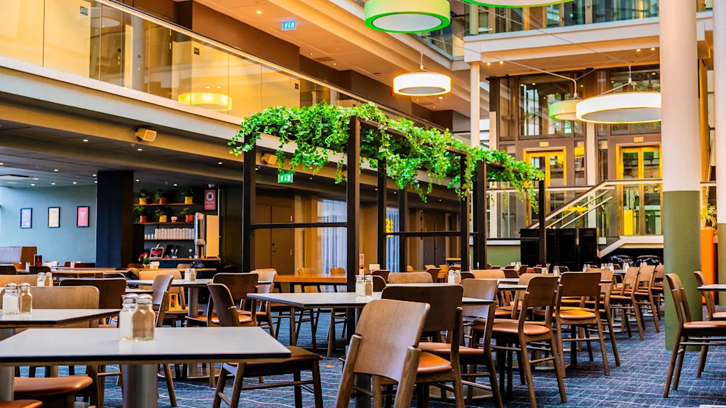 Ett rymligt hotellatrium med matbord, moderna cirkulära lampor och frodiga gröna växter, vilket skapar en inbjudande atmosfär.