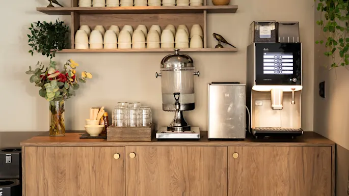 A self-service refreshment station with a coffee machine offering drink selection options, a water dispenser, and cups. PAPER bin.