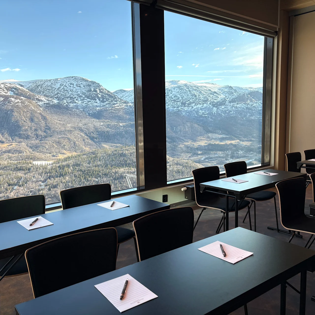 Desks with notebooks and pens are arranged in a classroom, set against a large window revealing a panoramic view of snowy mountains and a clear blue sky.