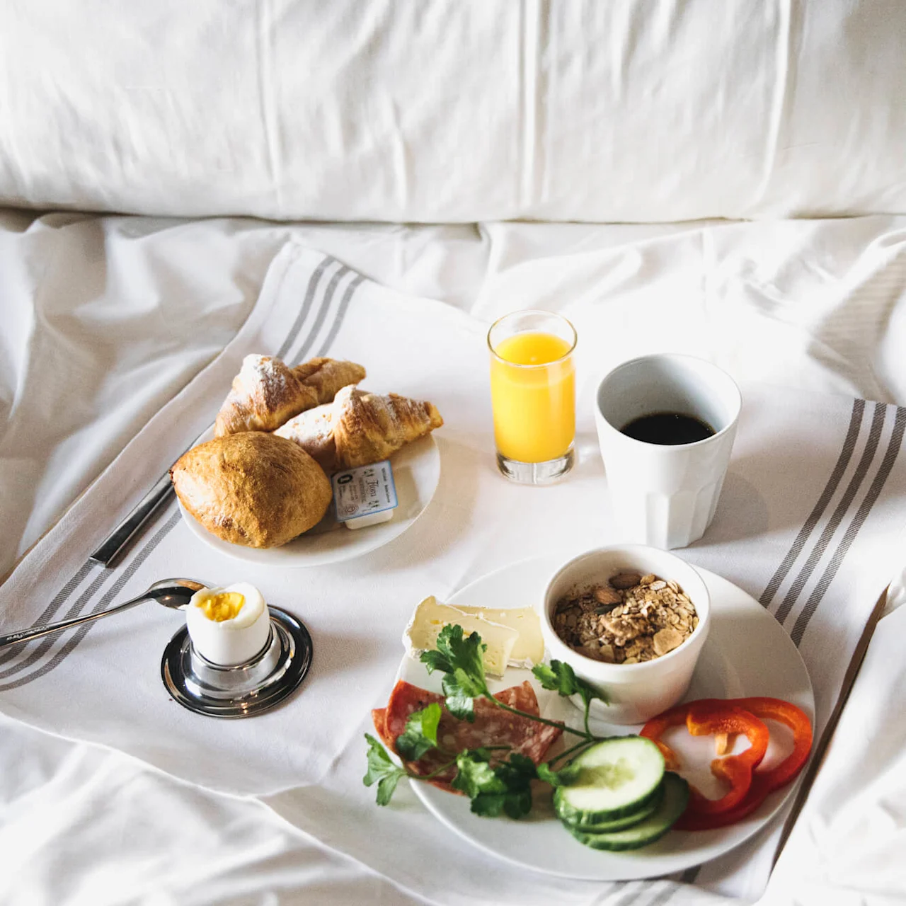 A breakfast tray holds a croissant, roll, butter, juice, coffee, soft-boiled egg, muesli, cheeses, salami, cucumber slices, and red pepper, resting on white bed sheets.