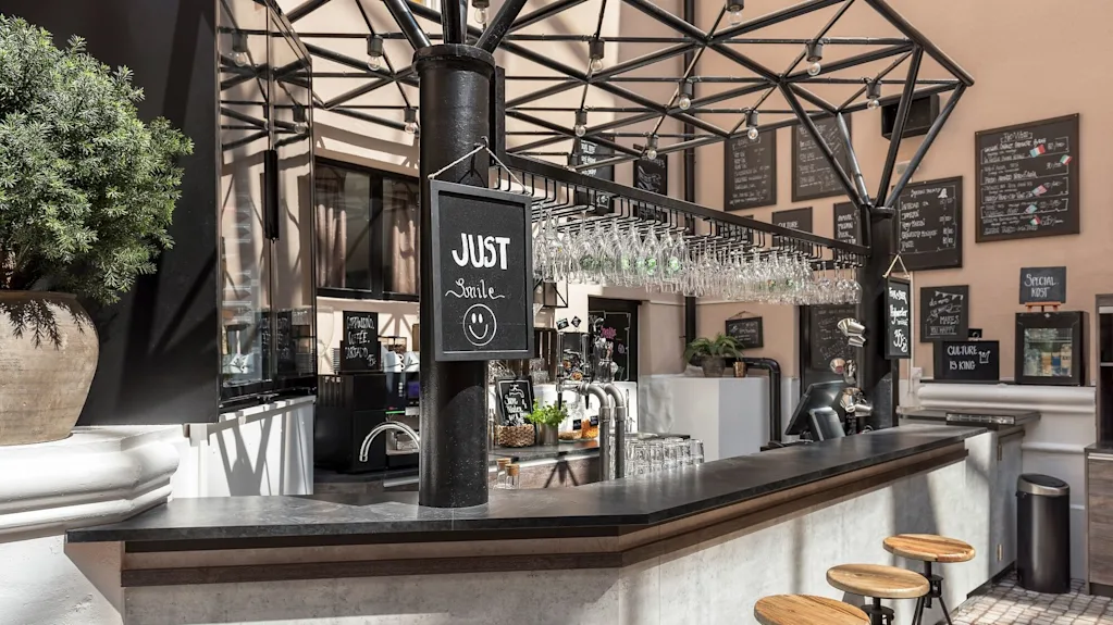 Outdoor bar with hanging glasses and sign saying "JUST smile" in a sunny, open-air courtyard with chalkboard menus.