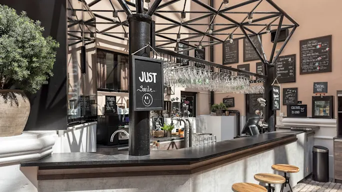 Utebar med hengende glass og skilt som sier "JUST smile" i en solfylt, åpen gårdsplass med krittavle-meny.