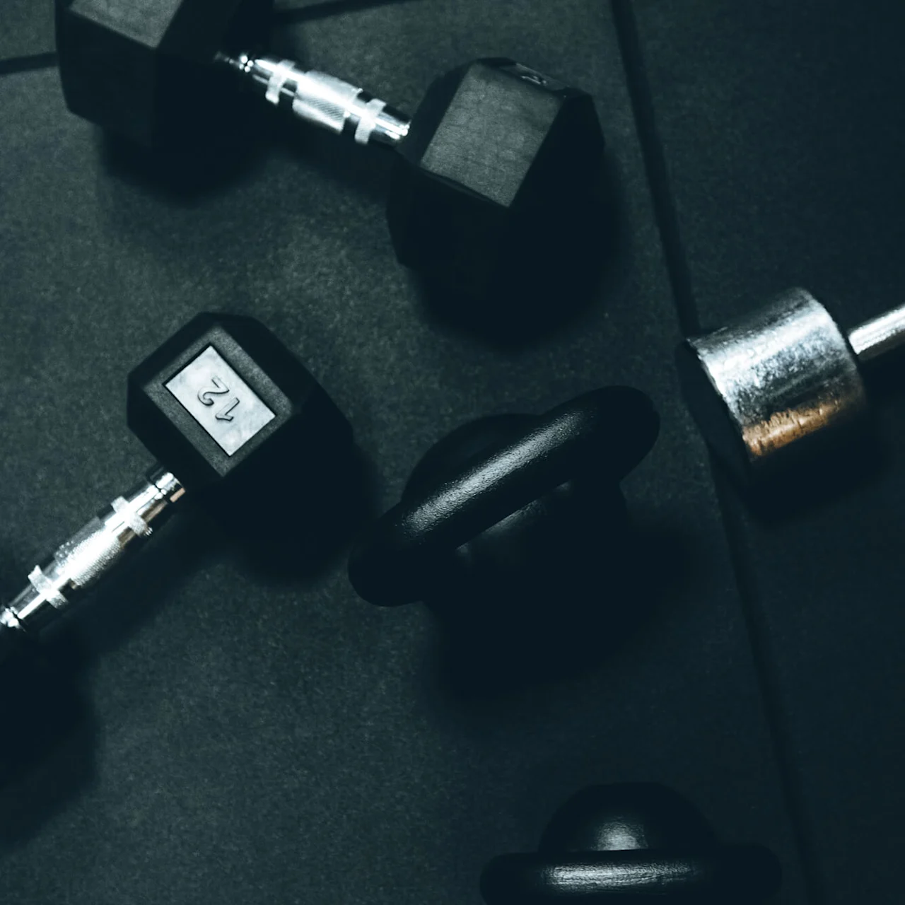 Dumbbells and kettlebells rest on a dark gym floor. One black dumbbell shows the number “12,” surrounded by other weights, creating a workout-ready environment.