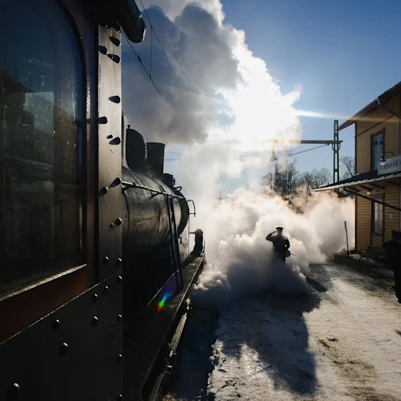 Ett ånglok på en snötäckt station, insvept i ett moln av vit ånga, med en person som går igenom det under en ljus himmel.