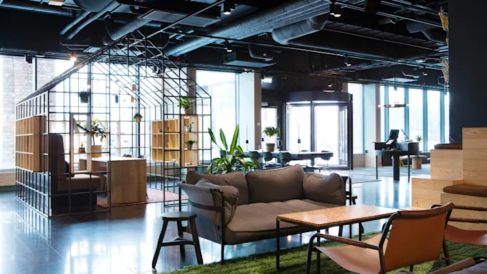 Et moderne åpent kontorlandskap med en blanding av uformelle sitteplasser og arbeidsstasjoner, store vinduer og et industrielt tak.