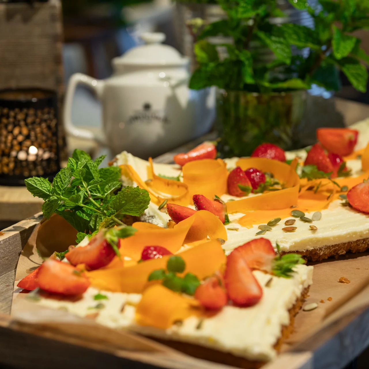 En rektangulær cheesecake pyntet med jordbær, mangostykker og frø ligger på en træbakke i hyggelige omgivelser. To tændte lys, en hvid tekande og en mynteplante står i nærheden.