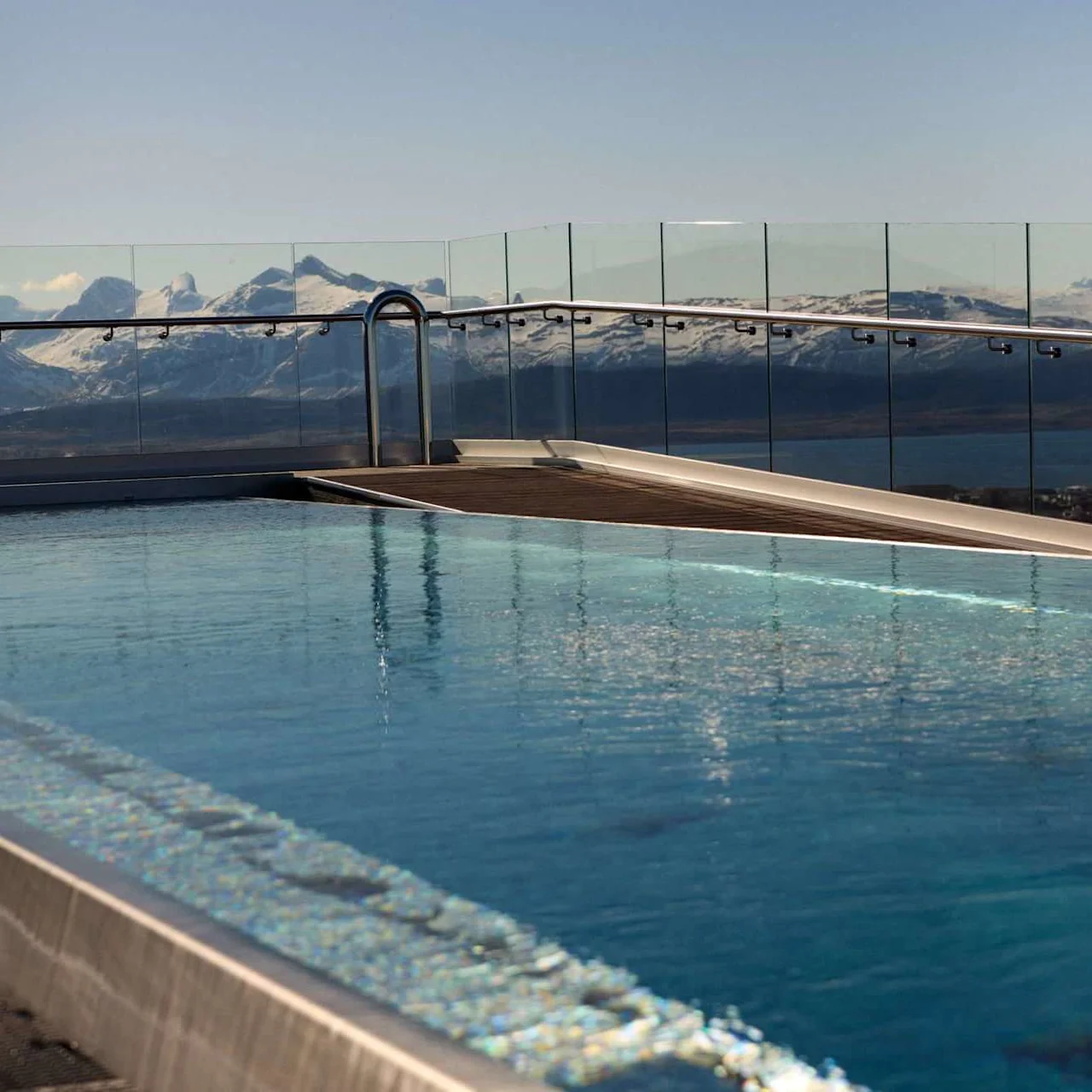 A rooftop swimming pool overlooks a serene mountain range with snow-capped peaks, surrounded by clear glass railings under a blue sky.