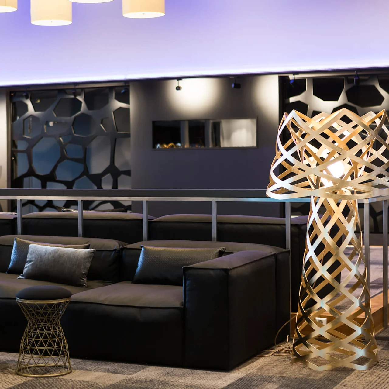 A modern lounge features a dark sofa, unique patterned lamp, and small tables, surrounded by abstract red sculptures on stands and decorative wall panels.