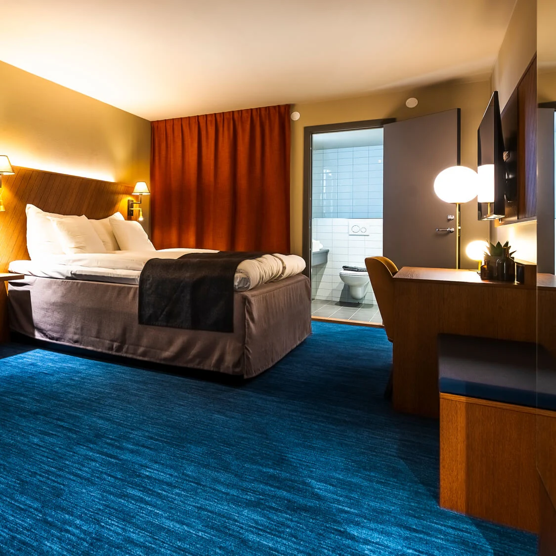 Ett modernt hotellrum med en väl bäddad säng, varmt ljus och en öppen badrumsdörr i en välutrustad miljö.