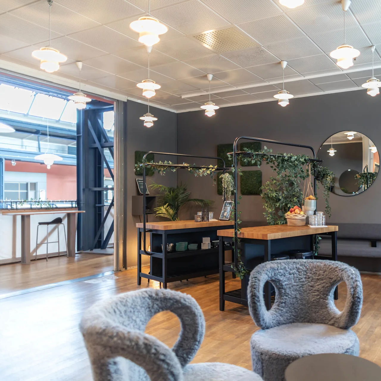 Cozy lounge with grey chairs and tables, adorned with hanging pendant lights and greenery. A large mirror decorates the grey wall, reflecting bright windows and adding depth.