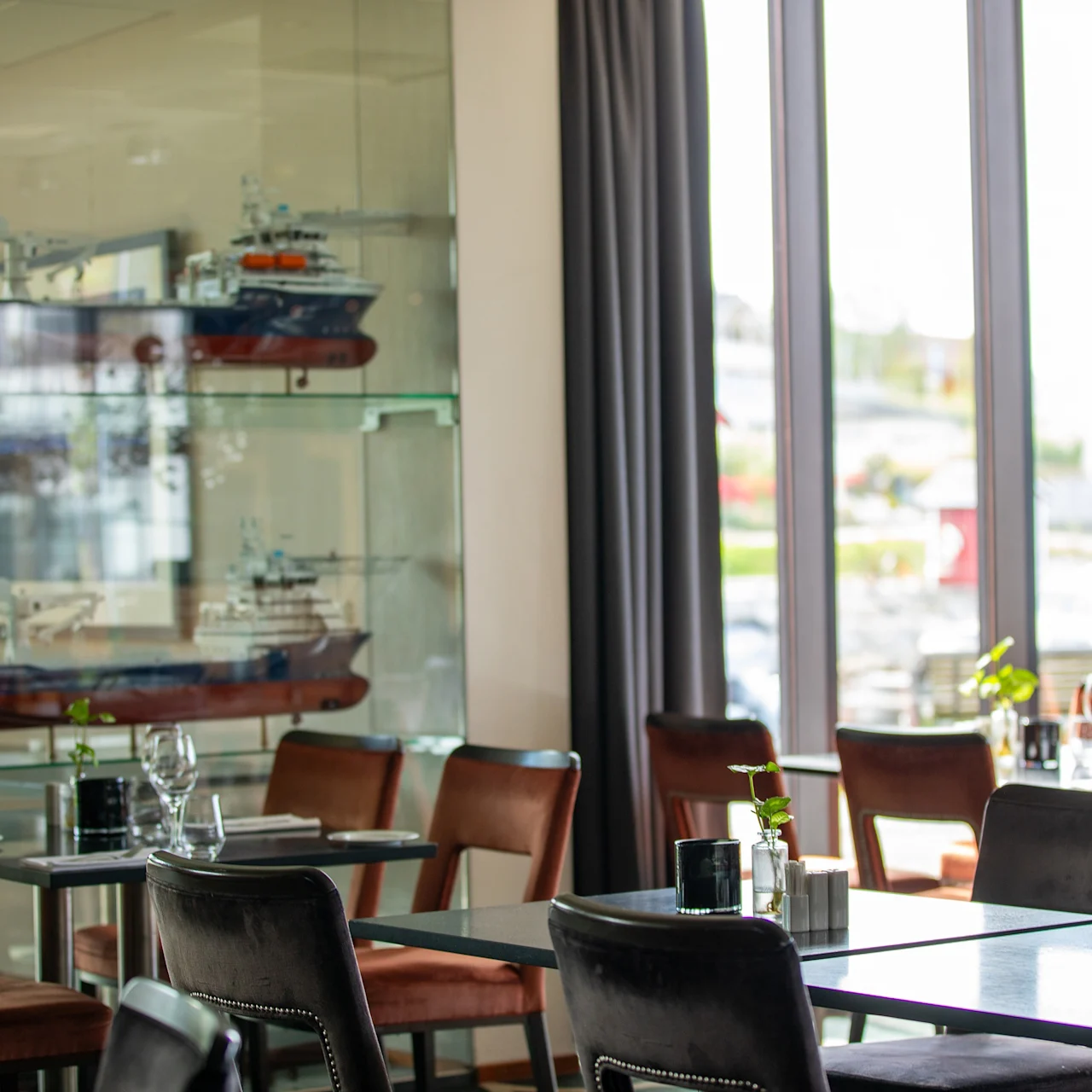 Modellskip vises i et glasskap, reflekterer lys i en solrik restaurant med pent oppstilte bord og stoler nær store vinduer. Ingen tekst er til stede.