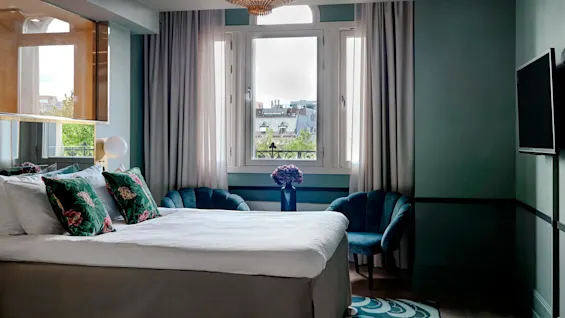 A bed with floral cushions stands in front of a window, flanked by blue chairs, beneath an elegant chandelier in a stylishly decorated bedroom.