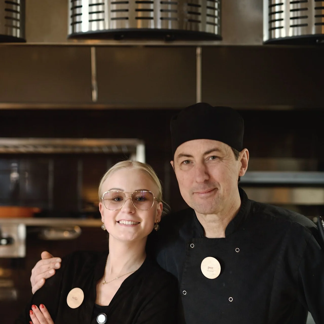 Two restaurant workers posing together with smiles, a woman in front and a man in chef attire behind, in a kitchen setting.