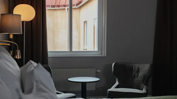 En säng med vita kuddar står nära två fåtöljer vid ett litet runt bord, upplyst av en lampa, i ett rum med ett fönster som visar en beige yttervägg.