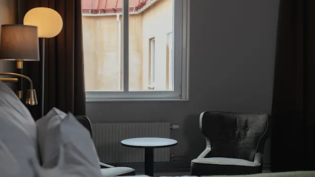 Bed with white pillows sits near two adjacent armchairs by a small round table, illuminated by a lamp, in a room with a window displaying an exterior beige wall.