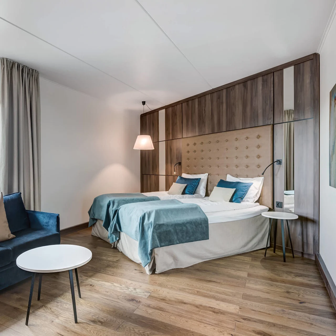 A cozy bedroom features two neatly made twin beds with blue and white bedding, a blue couch, and a small white round table. Wooden flooring and a large window provide a welcoming ambiance.